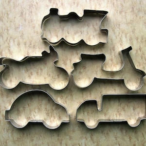 May include: Five metal cookie cutters shaped like different vehicles: a train, a motorcycle, a scooter, a car, and a truck. The cutters are on a brown background.