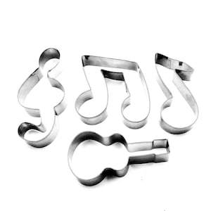Puede incluir: Un conjunto de cortadores de galletas de acero inoxidable con formas de notas musicales, una clave de sol y una guitarra. Los cortadores están dispuestos sobre un fondo blanco, ideales para hornear y para músicos.