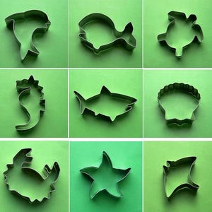 May include: Nine metal cookie cutters in various shapes, including a dolphin, fish, turtle, seahorse, shark, seashell, starfish, and stingray. The cutters are all silver in color and are arranged in three rows of three.