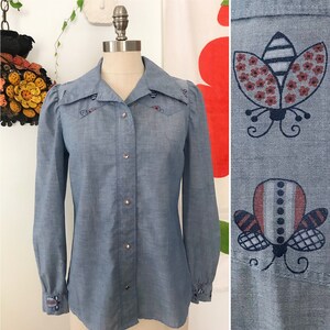May include: A light blue denim shirt with long sleeves and a button-down front. The shirt has embroidered butterflies on the collar, cuffs, and yoke. The butterflies are in shades of red, black, and white.