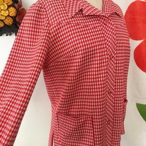 May include: A red and white houndstooth patterned jacket with a long sleeve, a collar, and a button closure. The jacket has a pocket on the left side.