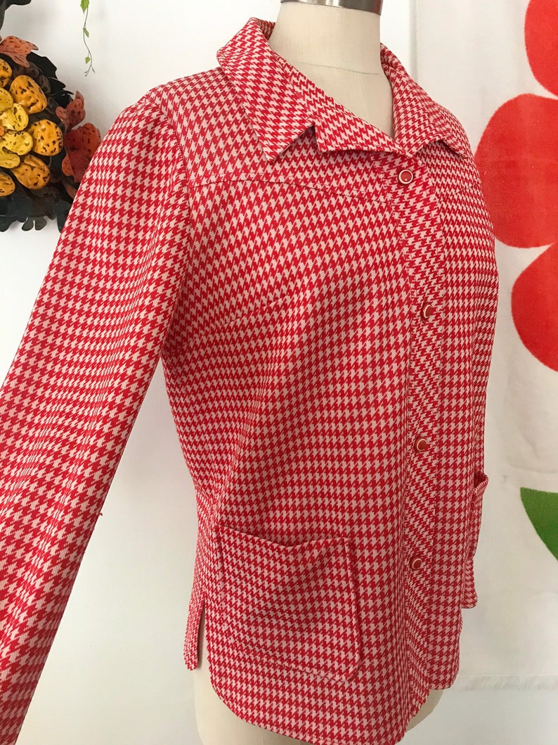 May include: A red and white houndstooth patterned jacket with a long sleeve, a collar, and a button closure. The jacket has a pocket on the left side.