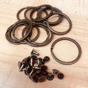 May include: A collection of bronze-colored key rings and rivets. The key rings are circular with a split for attaching keys. The rivets have a flat head and a post for securing materials. The items are arranged on a light wood surface.