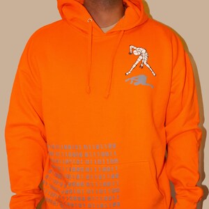May include: Orange hoodie with a reflective graphic of a stick figure doing a handstand. The hoodie has a large pocket on the front with a repeating pattern of binary code.