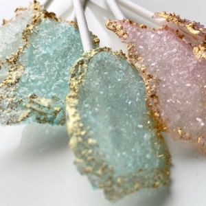 Puede incluir: Primer plano de varias piruletas de caramelo de roca en tonos azules, rosas y transparentes. Cada piruleta tiene un borde dorado y está montada en un palito blanco. El caramelo tiene una textura cristalina.