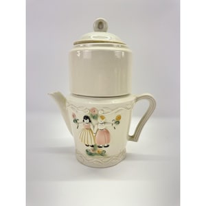May include: A cream-colored ceramic teapot with a matching lid, featuring a raised relief design of two figures holding flowers. The figures are painted with black hair, a yellow skirt, and a pink skirt, surrounded by small floral accents.