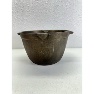 May include: A dark, weathered metal mixing bowl with a rounded base and a wide, open top. The bowl features two small, protruding side handles and a small pouring spout integrated into the rim, showing a tarnished, aged patina throughout.