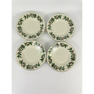 May include: Four round off-white saucers arranged in a two-by-two grid. Each saucer features a decorative border pattern consisting of green holly leaves and small red berries encircling the center. The edges of the saucers are smooth and circular.