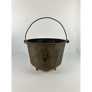 May include: A dark, weathered cast iron cauldron featuring a rounded body, three short legs, and a thin, arched metal handle. The exterior surface displays a mottled, rusted patina with a single metal ring attached to the front side of the vessel.