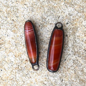 May include: Two elongated, reddish-brown pendants with a striped pattern and a dark metal frame. Each pendant has a small loop at the top. The pendants are displayed on a textured, light-colored surface.