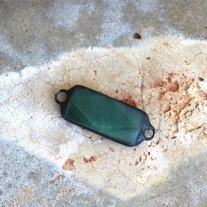 May include: A rectangular green glass cabochon set in a dark metal frame with a small circular loop on each end. The glass has a smooth, polished surface with subtle internal variations and a deep, translucent emerald green color throughout.