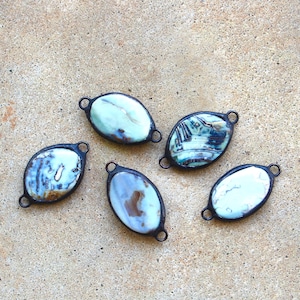 May include: Five oval-shaped pendants with a marbled, blue-green design set in a dark metal frame. Each pendant has a small loop at the top for attaching to a chain or other jewelry component. The pendants are displayed on a textured, tan surface.