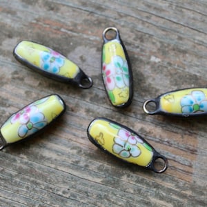 May include: Five yellow ceramic beads with floral designs and black metal accents. Each bead has a loop at the top for attaching to a necklace or bracelet.