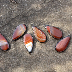 May include: Six teardrop-shaped agate pendants with a silver wire wrap. The agate is a mix of brown, orange, and white.