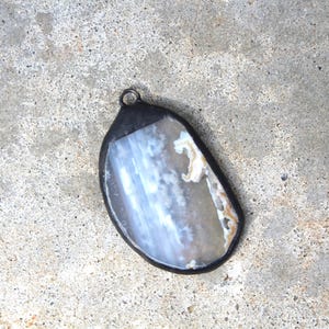 May include: A pendant with a natural stone center, featuring shades of white, gray, and brown. The stone is encased in a dark, possibly black, metal frame with a small loop at the top for a chain. The pendant is set against a textured gray surface.