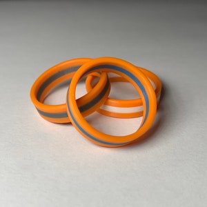 May include: Three orange silicone rings with a contrasting stripe in gray, white, and blue. The rings are stacked and slightly overlapping, showcasing their design. The rings are a vibrant accessory.