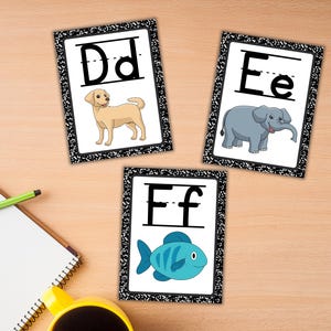 Może przedstawiać: Trzy edukacyjne fiszki z literami D, E i F. Każda karta ma czarno-białą obw&oacute;dkę i odpowiadającą ilustrację zwierzęcia: psa, słonia i ryby. Karty leżą na drewnianej powierzchni.
