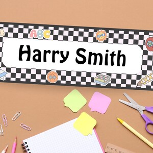 May include: A personalized name tag with the name "Harry Smith" in bold black letters. The tag has a black and white checkered border and is decorated with colorful school-themed stickers, including an apple, pencil, and ABC letters. School supplies are scattered around the name tag.