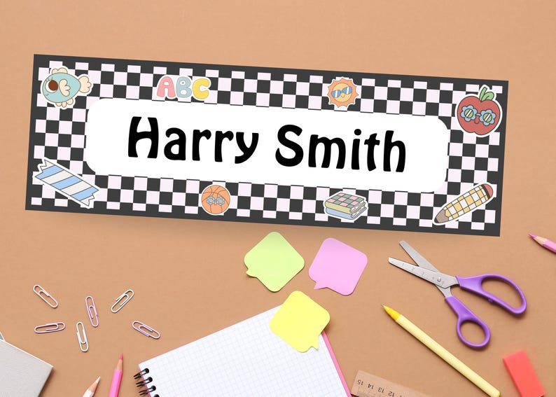 May include: A personalized name tag with the name "Harry Smith" in bold black letters. The tag has a black and white checkered border and is decorated with colorful school-themed stickers, including an apple, pencil, and ABC letters. School supplies are scattered around the name tag.