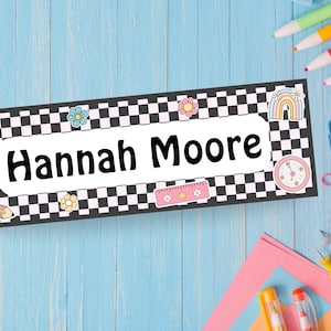 May include: A rectangular name tag with the name "Hannah Moore" in bold black letters. The tag has a black and white checkered border and is decorated with colorful illustrations of flowers, a rainbow, an apple, a pencil, and a ruler.