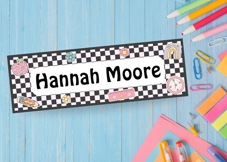 May include: A rectangular name tag with the name "Hannah Moore" in bold black letters. The tag has a black and white checkered border and is decorated with colorful illustrations of flowers, a rainbow, an apple, a pencil, and a ruler.