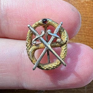 May include: A gold-toned brooch featuring a textured, circular snake design with two crossed silver-toned swords layered over the center. A small, dark red gemstone is set at the top of the snake's head, completing the detailed metal pin.