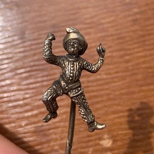 Puede incluir: Un alfiler de palo plateado con una figura detallada de un hombre con un sombrero y ropa adornada. El hombre está representado en una pose de baile con los brazos en alto.