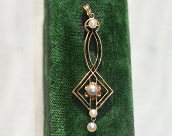 Op de afbeelding: Een gouden hanger met een geometrisch ontwerp en drie parels die eraan hangen. De hanger wordt gepresenteerd op een groene fluwelen achtergrond.