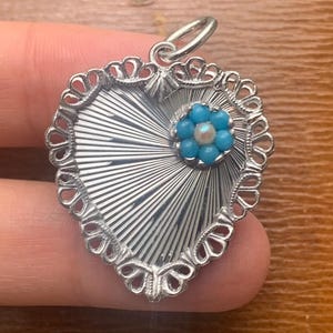 May include: Silver heart-shaped pendant with a detailed, rayed design. The pendant features a delicate, openwork border and a small flower with turquoise beads and a central pearl. A small loop is at the top for a chain.