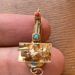Op de afbeelding: Een goudkleurige bedel met een liggende figuur op een rechthoekige basis, versierd met rode en turkooizen edelstenen. De bedel heeft een kleine lus voor bevestiging. De bedel wordt door een persoon vastgehouden.