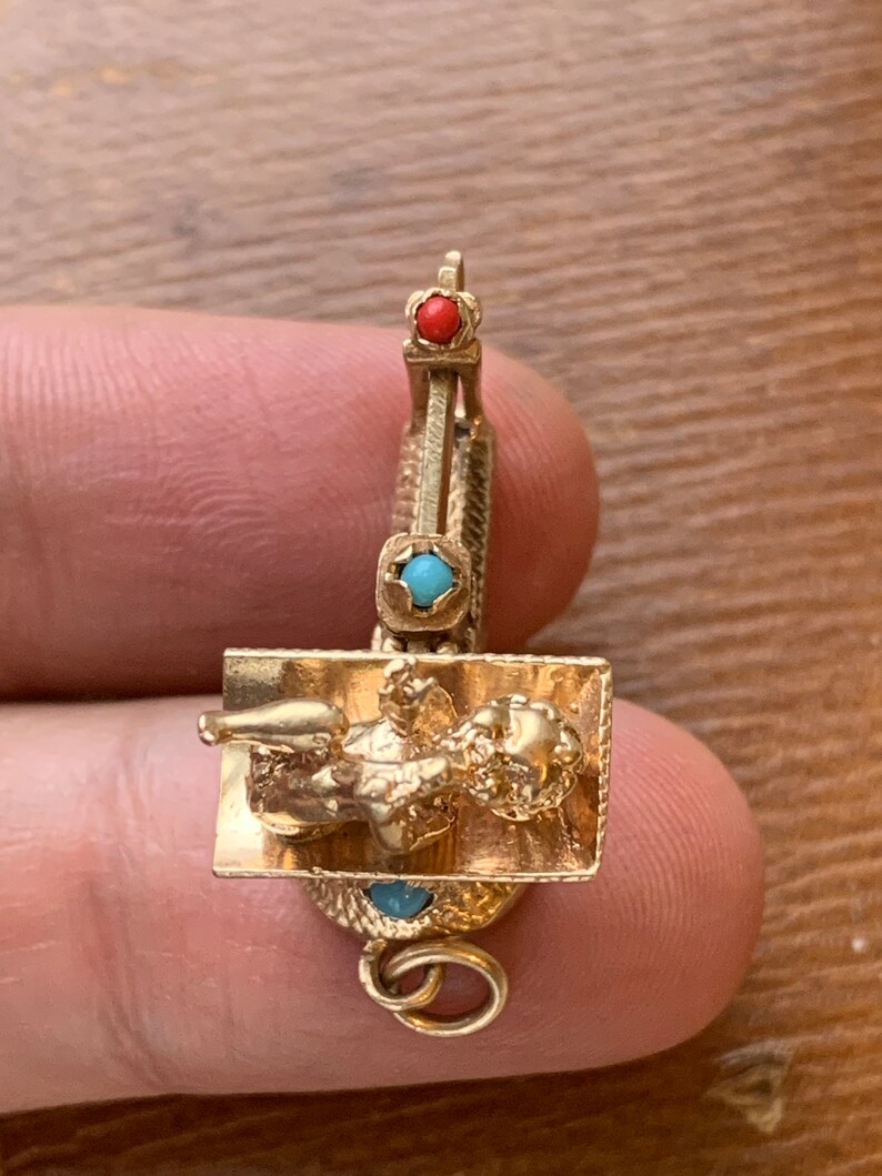 Op de afbeelding: Een goudkleurige bedel met een liggende figuur op een rechthoekige basis, versierd met rode en turkooizen edelstenen. De bedel heeft een kleine lus voor bevestiging. De bedel wordt door een persoon vastgehouden.