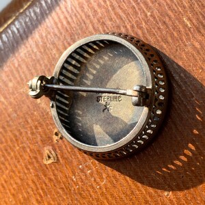 K&ouml;nnte beinhalten: Eine silberne Brosche mit einem filigranen Design und einer runden, silberfarbenen Mitte. Die R&uuml;ckseite der Brosche hat eine Nadel und eine Markierung mit der Aufschrift "STERLING" mit einem Stern.