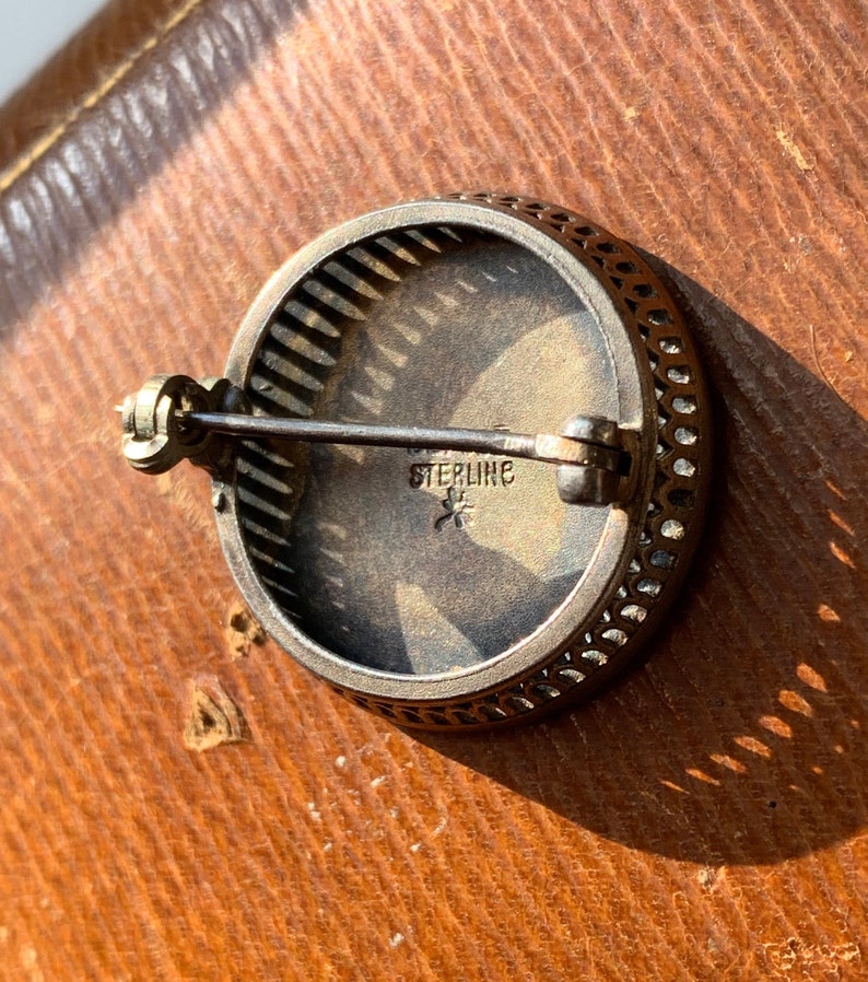 K&ouml;nnte beinhalten: Eine silberne Brosche mit einem filigranen Design und einer runden, silberfarbenen Mitte. Die R&uuml;ckseite der Brosche hat eine Nadel und eine Markierung mit der Aufschrift "STERLING" mit einem Stern.