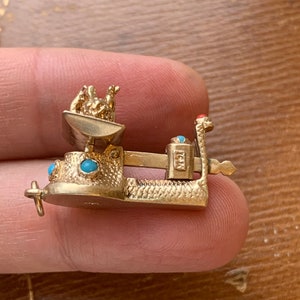Op de afbeelding: Een goudkleurige miniatuur weegschaal charm met turquoise en rode accenten. De weegschaal heeft een kleine kom met figuren, een balansbalk en een basis. De bedel wordt tussen vingers gehouden.