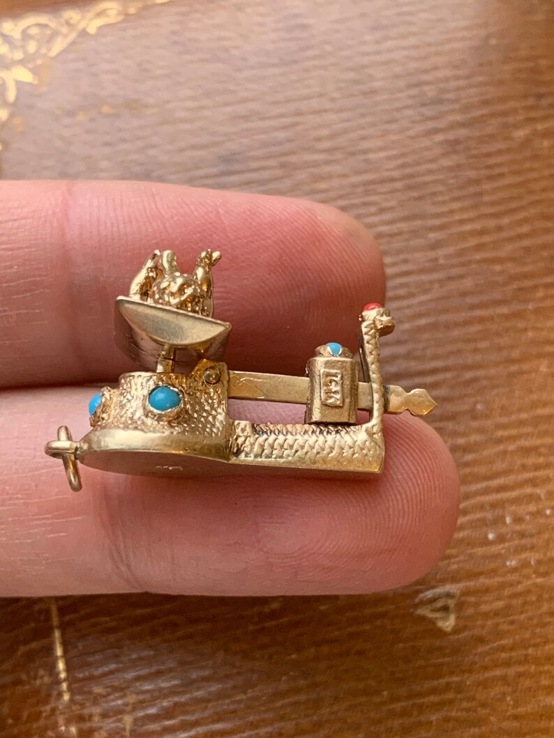 Op de afbeelding: Een goudkleurige miniatuur weegschaal charm met turquoise en rode accenten. De weegschaal heeft een kleine kom met figuren, een balansbalk en een basis. De bedel wordt tussen vingers gehouden.
