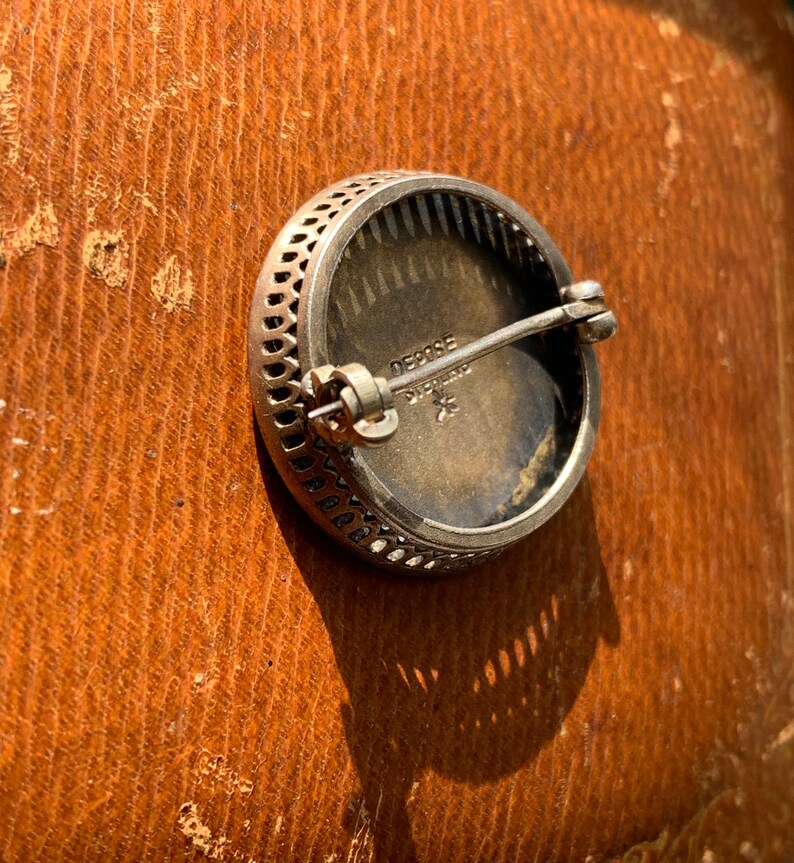 K&ouml;nnte beinhalten: Eine silberne Brosche mit einem dekorativen Filigranmuster am Rand. Die R&uuml;ckseite der Brosche tr&auml;gt den Text "DECOBE STERLING" und einen Stern.