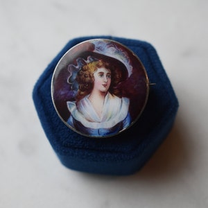 K&ouml;nnte beinhalten: Eine silberne Brosche mit einem gemalten Portr&auml;t einer Frau in einem wei&szlig;en Hut und einem blauen Kleid. Das Portr&auml;t ist vor einem dunkelvioletten Hintergrund platziert.