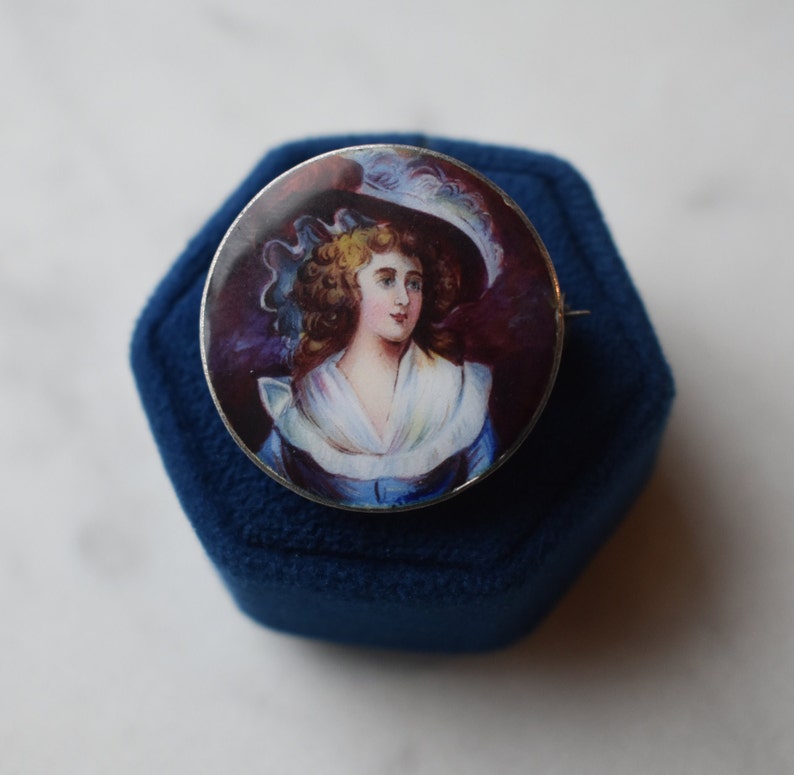 K&ouml;nnte beinhalten: Eine silberne Brosche mit einem gemalten Portr&auml;t einer Frau in einem wei&szlig;en Hut und einem blauen Kleid. Das Portr&auml;t ist vor einem dunkelvioletten Hintergrund platziert.
