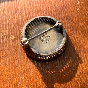 K&ouml;nnte beinhalten: Eine runde silberne Brosche mit einem dekorativen Filigranrand. Die R&uuml;ckseite der Brosche tr&auml;gt die Inschrift "Depose Sterling" und einen Stern.