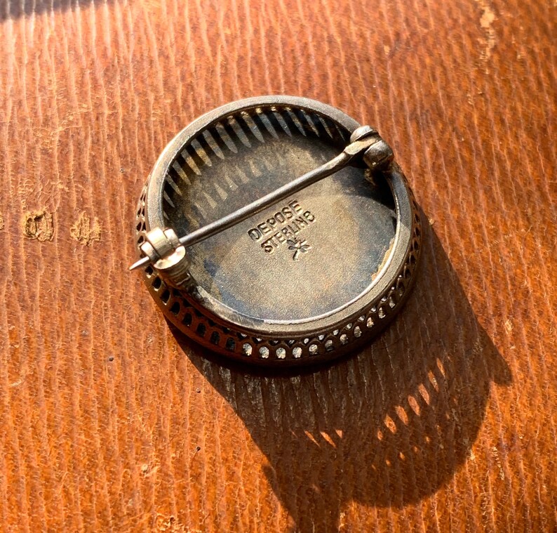 K&ouml;nnte beinhalten: Eine runde silberne Brosche mit einem dekorativen Filigranrand. Die R&uuml;ckseite der Brosche tr&auml;gt die Inschrift "Depose Sterling" und einen Stern.
