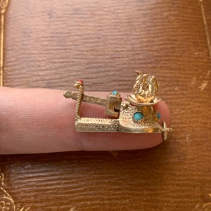 Op de afbeelding: Een goudkleurige bedel met turquoise en rode accenten. De bedel heeft een gedetailleerd ontwerp met een kleine vogel figuur en een decoratieve basis. De bedel wordt door een vinger vastgehouden.