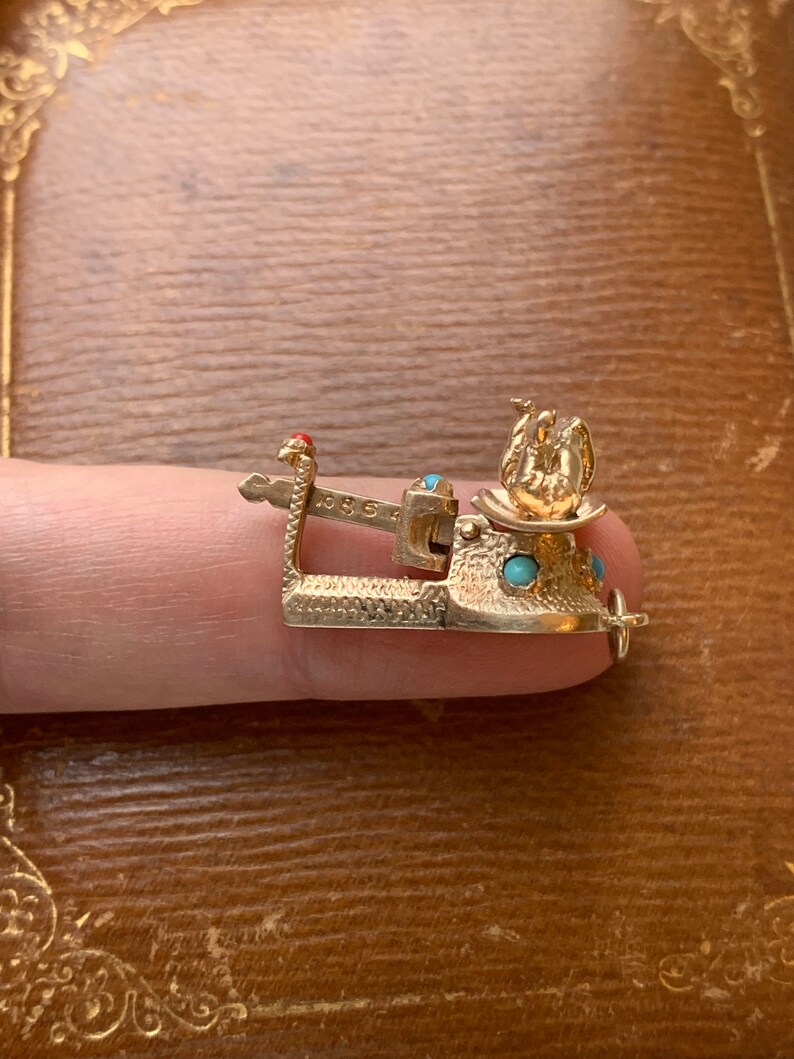 Op de afbeelding: Een goudkleurige bedel met turquoise en rode accenten. De bedel heeft een gedetailleerd ontwerp met een kleine vogel figuur en een decoratieve basis. De bedel wordt door een vinger vastgehouden.