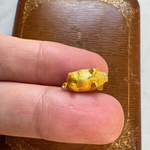 May include: A small, gold-colored pig figurine. The pig is held between two fingers, with a brown, decorative box in the background. The pig is detailed with visible ears, snout, and legs. The box has a decorative, embossed design.