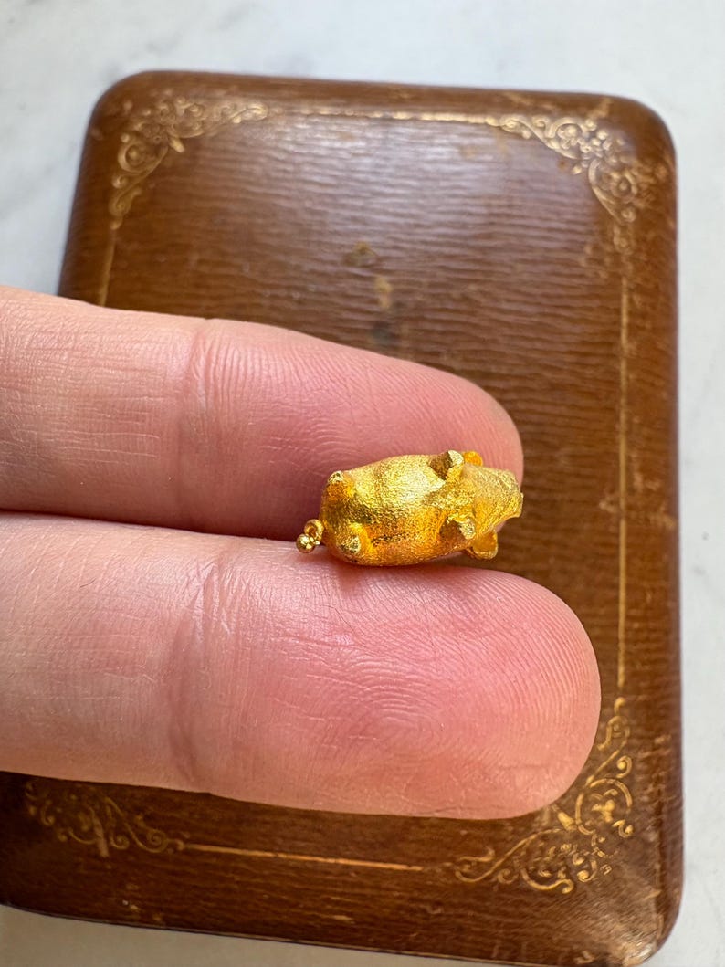 May include: A small, gold-colored pig figurine. The pig is held between two fingers, with a brown, decorative box in the background. The pig is detailed with visible ears, snout, and legs. The box has a decorative, embossed design.