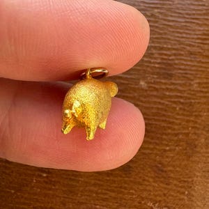 May include: A small, gold-colored pig charm with a textured surface. The charm is held between two fingers, showcasing its three-dimensional design and a small loop for attachment. The pig has four legs, a tail, and small ears.