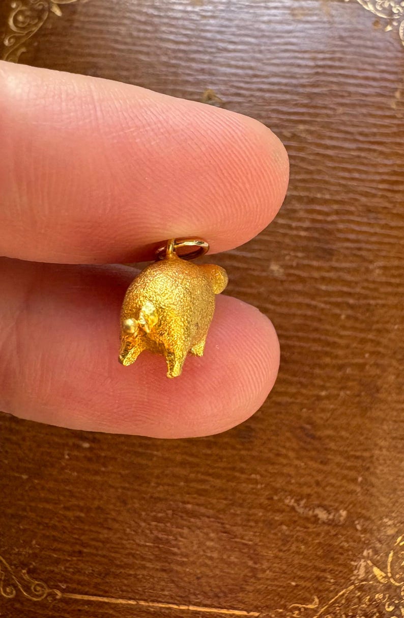 May include: A small, gold-colored pig charm with a textured surface. The charm is held between two fingers, showcasing its three-dimensional design and a small loop for attachment. The pig has four legs, a tail, and small ears.