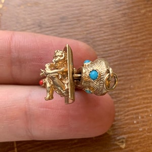 Op de afbeelding: Een goudkleurige bedel met een zittende cherubijn die fluit speelt. De bedel heeft een gestructureerd, bolvormig element versierd met kleine turquoisekleurige stenen. Een klein rood accent is zichtbaar.