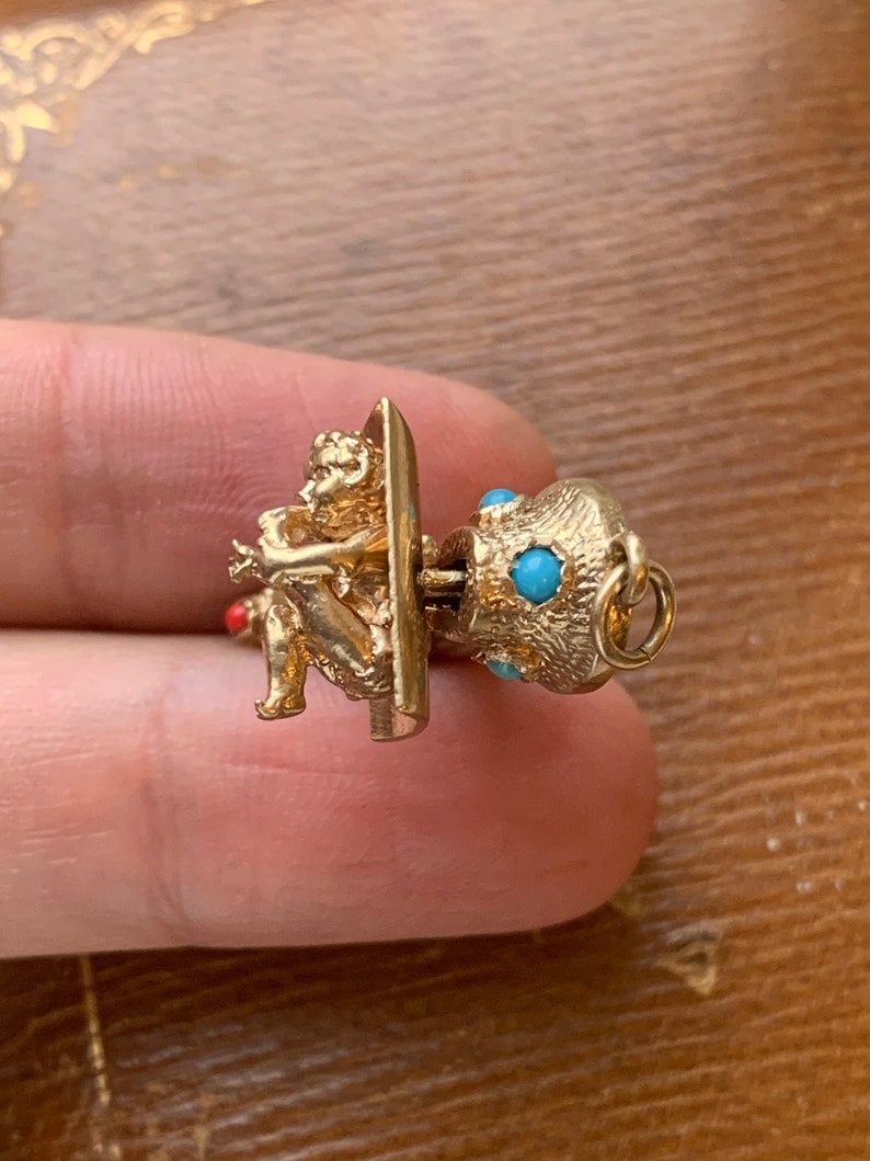 Op de afbeelding: Een goudkleurige bedel met een zittende cherubijn die fluit speelt. De bedel heeft een gestructureerd, bolvormig element versierd met kleine turquoisekleurige stenen. Een klein rood accent is zichtbaar.