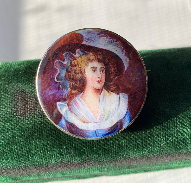 K&ouml;nnte beinhalten: Eine runde silberne Brosche mit einem gemalten Portr&auml;t einer Frau in einem blauen Kleid und einem wei&szlig;en Hut mit Federn. Der Hintergrund ist dunkelviolett.