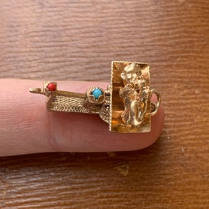 Op de afbeelding: Een goudkleurige bedel met een rechthoekig frame met een gebeeldhouwde figuur. De bedel heeft een kleine rode en turkooizen steen. De bedel zit op een vinger.